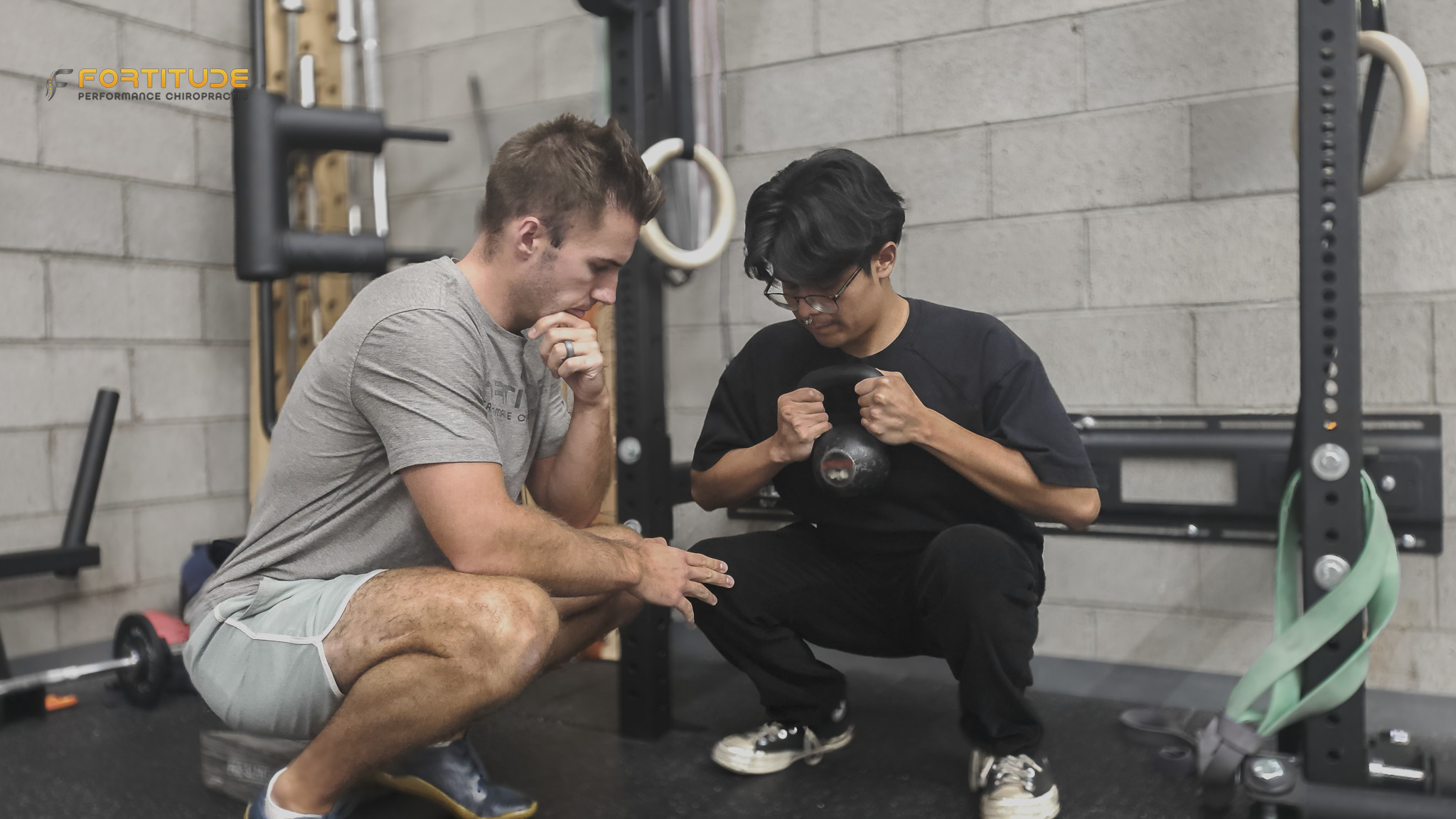 A coach kneels next to a client on a bench, showing something on a phone screen. Both appear focused and engaged. The setting is a well-equipped gym with racks, cables, and resistance bands in the background.
