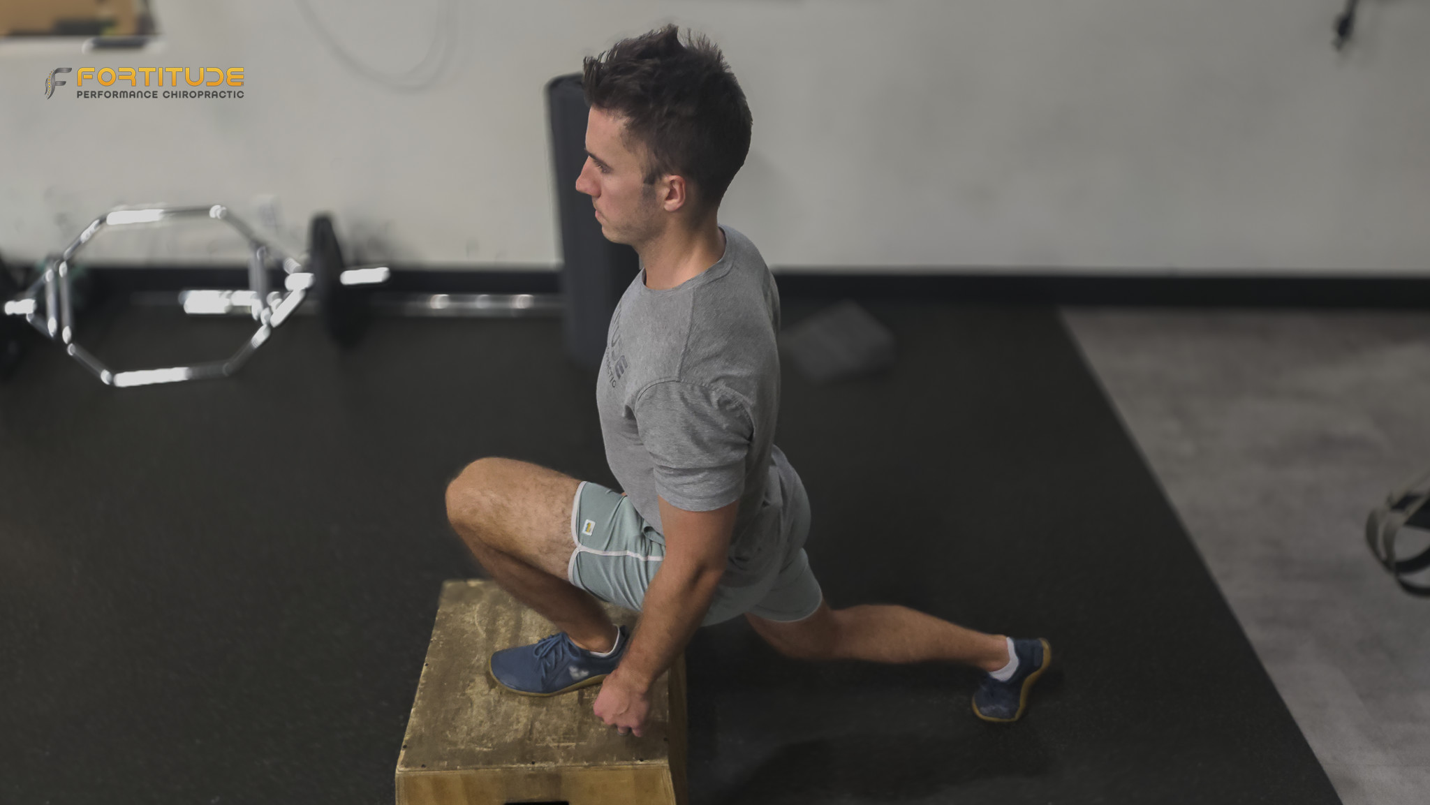 The athlete holds a dumbbell while positioned in a lunge, with a resistance band providing tension from behind. Natural light enters through the window, and gym equipment lines the wall nearby.