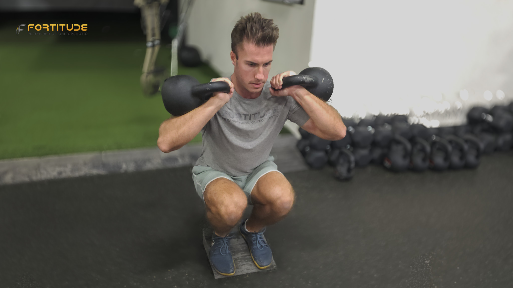 A man in a gray athletic outfit performs a squat while holding two kettlebells at shoulder height. His heels are elevated on slant boards, emphasizing ankle mobility and quad engagement. A turf strip and kettlebell rack are visible in the background.