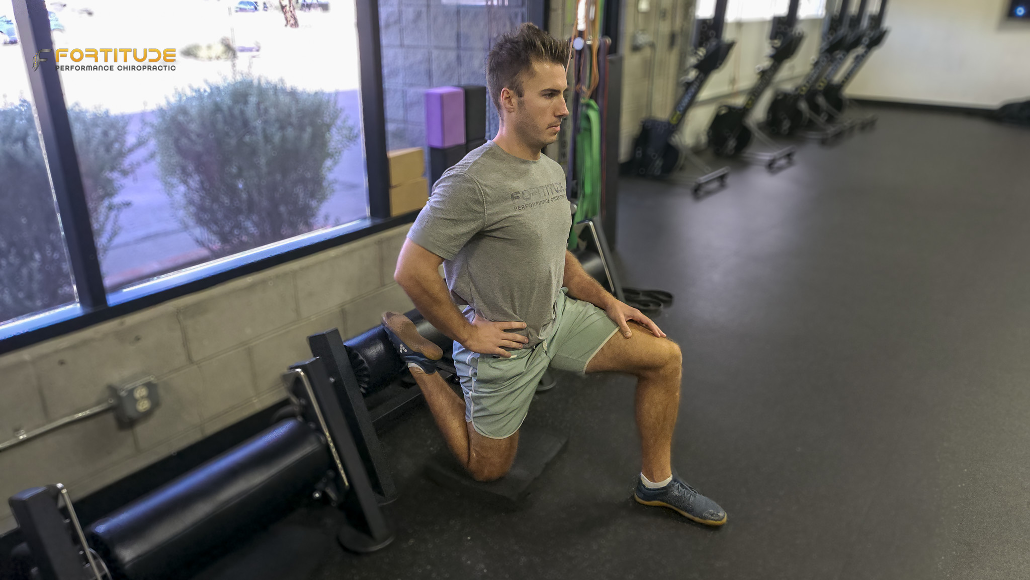 The athlete executes a Bulgarian split squat with his rear foot elevated on a foam pad and his front leg bent at 90 degrees. He is looking forward, maintaining posture and balance. The setup is in a gym corner with a squat rack behind him.